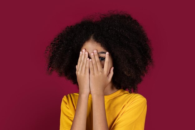 Terrified kid looking through fingers isolated on burgundy studio background