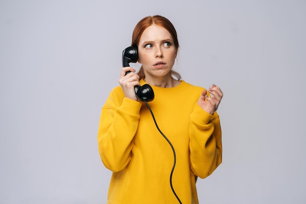 Terrified scared young woman wearing stylish yellow sweater talking on retro phone looking away