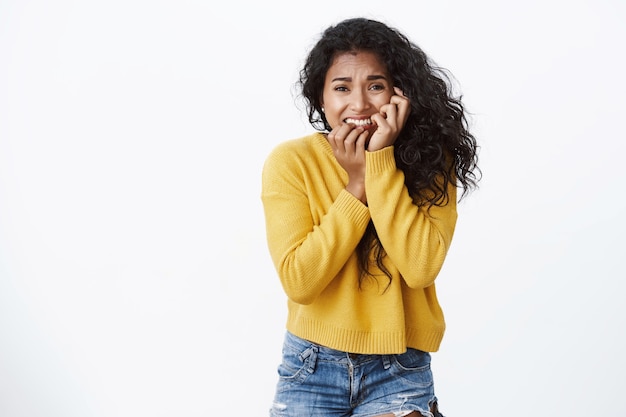 Terrified, scared young woman in yellow sweater, biting fingers, frowning and touching face from fear, stopping afraid stare camera frightened, standing white wall