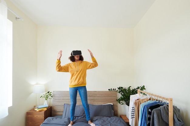 Terrified teenage mixed race girl in yellow sweater standing with hands up on bed while playing interactive video game using virtual reality device