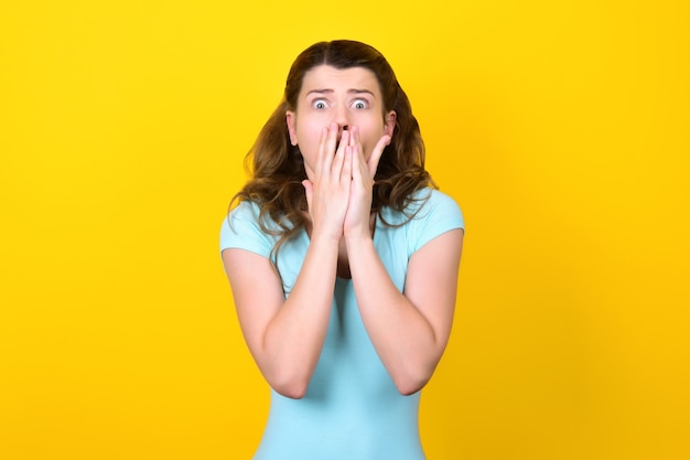 Terrified young woman with bulging eyes covers her mouth with her hands.