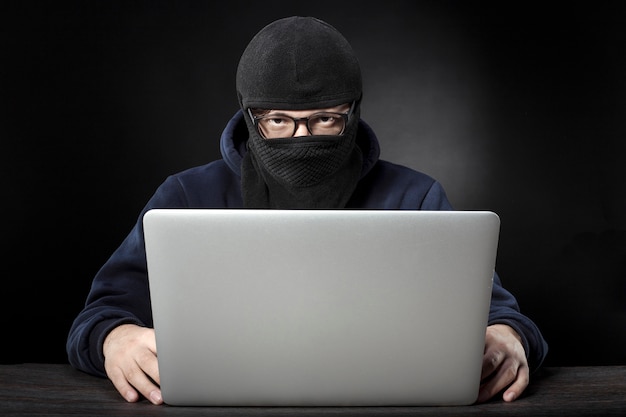 Terrorist in mask and glasses sitting at a laptop on a dark wall