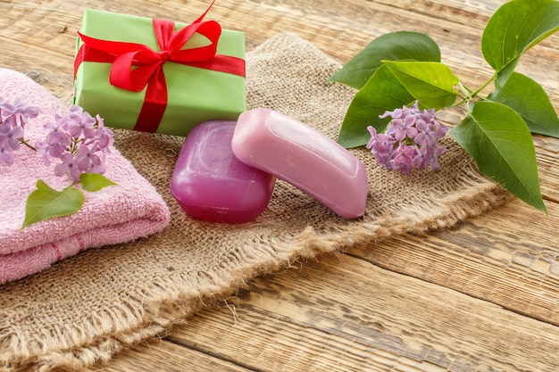 Asciugamano di spugna, sapone per le procedure del bagno, confezione regalo e fiori lilla su tela di sacco e assi di legno. prodotti e accessori termali.