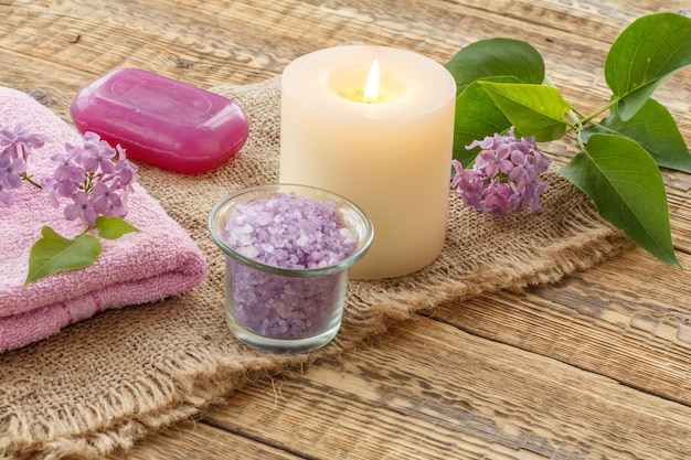 Terry towel, soap for bathroom procedures, sea salt, burning candle and lilac flowers on sackcloth and old wooden boards. Spa products and accessories.