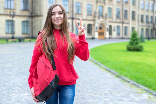 Terug naar school-concept. portret van een in lichte jeans vrijetijdskleding jongere duizendjarige dame geven v-sign staande in de buurt van deuren van educatief gebouw