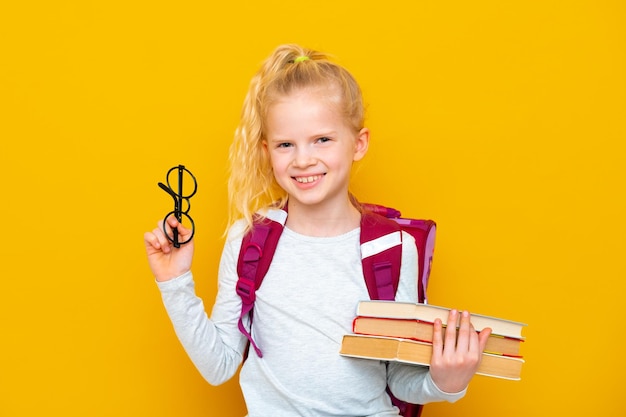 Terug naar school Portret van blond schoolmeisje in ronde bril met tas en boeken Gele studio achtergrond Onderwijs glimlachend in de camera