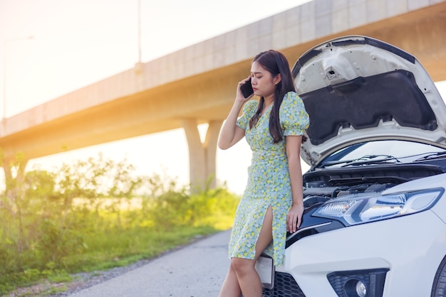 Terwijl ze naar een kapotte auto op straat kijkt, zit een jonge vrouw op haar telefoon. Bel een autoreparatie met een mobiele telefoon als de auto kapot is.