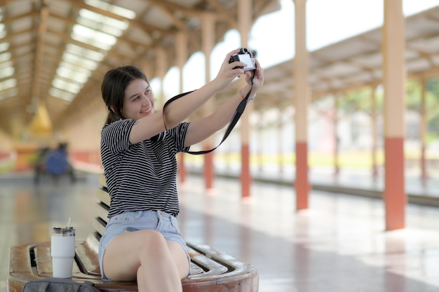 Terwijl ze op een reis wacht, gebruikt een jonge vrouwelijke toerist een selfiecamera, nemen buitenlandse adolescente meisjes selfies, wordt een witte reisbeker op de stoel geplaatst en wordt een reisreis gemaakt.