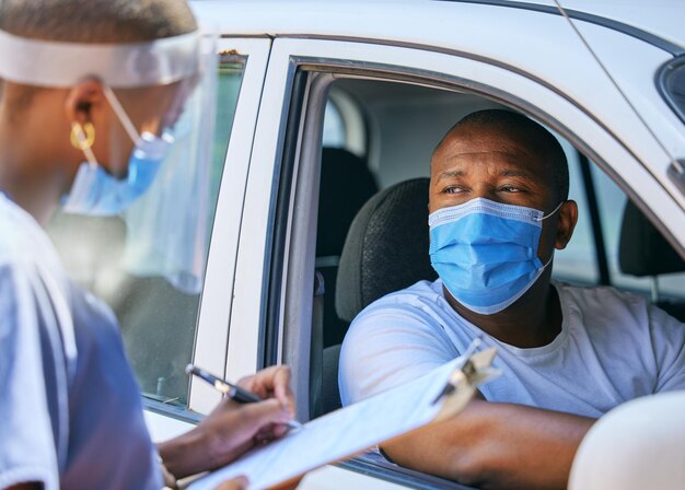 チェックポイントを通過するcovidドライブでのテストの質問とスクリーニングフェイスマスクを着用してコロナウイルスの詳細を書いている医療専門家と話している車で旅行している男性