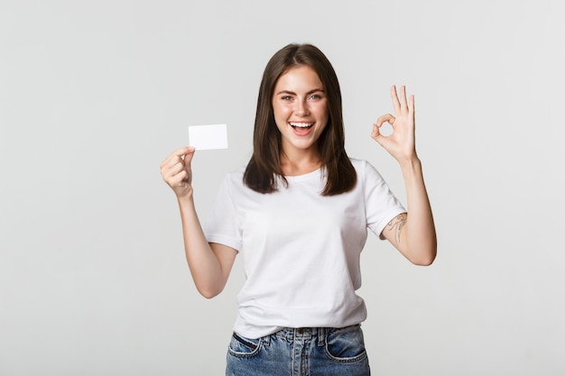 Tevreden aantrekkelijk donkerbruin meisje dat creditcard en ok gebaar toont, raadt bank aan.