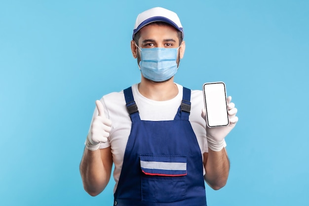 Tevreden adviseur in overall, masker en handschoenen met mobiele telefoon met mock-up display en duimen omhoog. Goede app voor online bezorgbestelling, onderhoudsdiensten voor huisreparatie. studio-opname binnenshuis