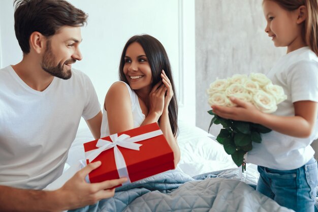 Tevreden brunette vrouw die op bed zit en naar haar knappe man kijkt terwijl haar schattige dochter bloemen vasthoudt