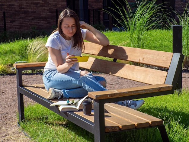 Tevreden brunette vrouw zittend op een bankje en online studeren met smartphone in park Onderwijs school en mensen concept Vrouwelijke student studeert op bankje op campus Meisje zit met gele telefoon