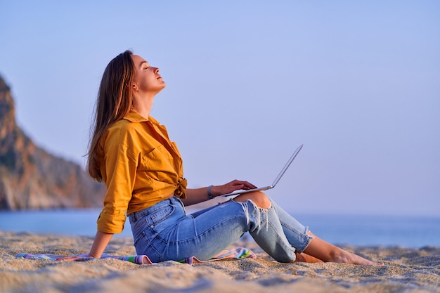 Tevreden duizendjarige gratis freelancer-vrouw die computer gebruikt en op het zandstrand aan zee zit genot van droomkantoorconcept voor werken op afstand