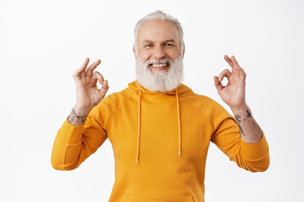 Tevreden en gelukkige senior man met tatoeages toont oké ok-tekens, knikt goedkeurend, lacht en glimlacht tevreden, prees uitstekend werk, goed gedaan gebaar, maak compliment niet slecht