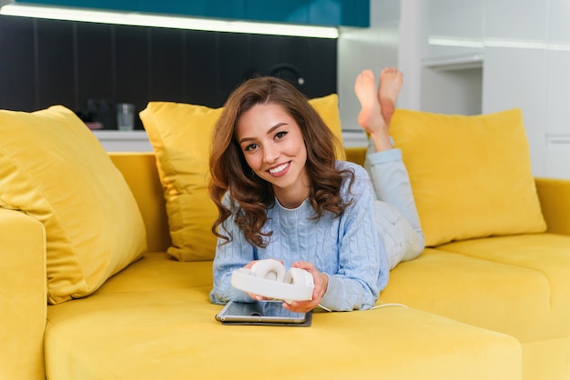 Tevreden gelukkig meisje met behulp van tablet terwijl liggend op comfortabele gele bank en genieten van aangename deuntjes met koptelefoon.