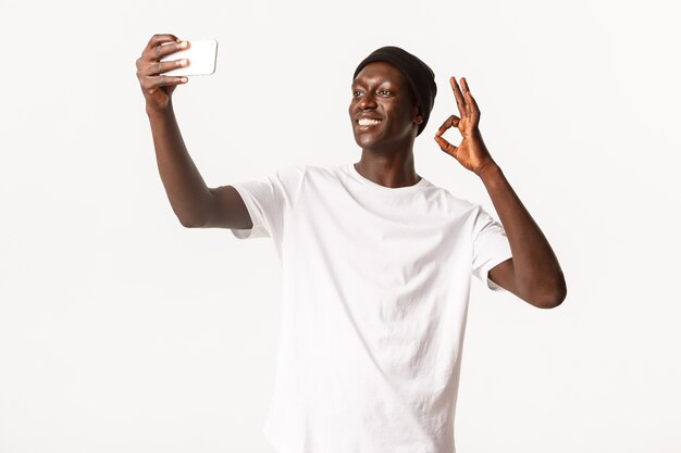 Tevreden, glimlachende Afro-Amerikaanse jonge kerel in muts, selfie of videogesprek via smartphone nemen, ok gebaar tonen op zelfportret