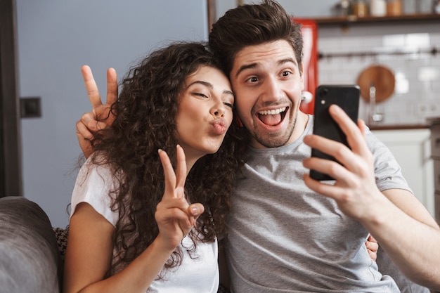 tevreden jong stel man en vrouw die thuis op de bank zitten en samen selfie-foto maken op smartphone