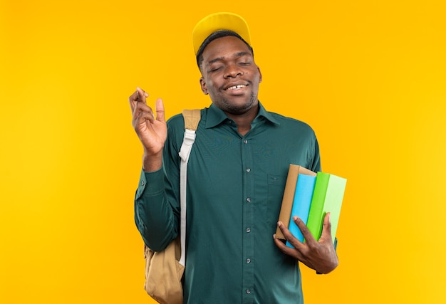 Tevreden jonge Afro-Amerikaanse student met pet en rugzak die boeken vasthoudt en zijn vingers kruist geïsoleerd op een oranje muur met kopieerruimte