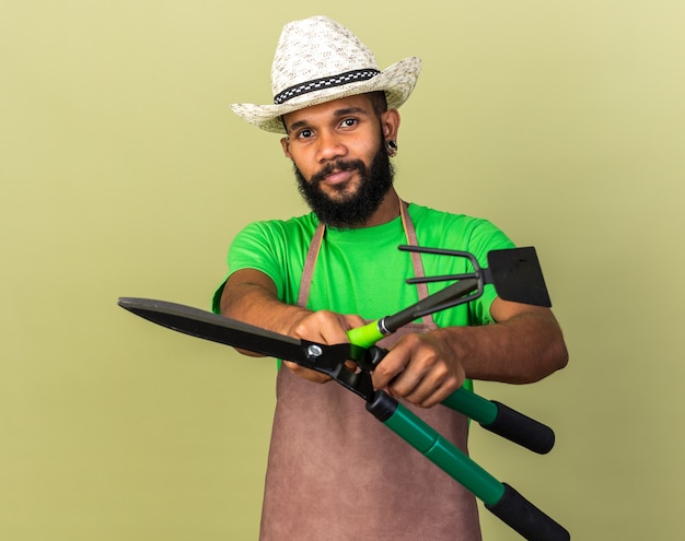 Tevreden jonge afro-amerikaanse tuinman met een tuinhoed die een tondeuse met een schoffelhark op de camera vasthoudt