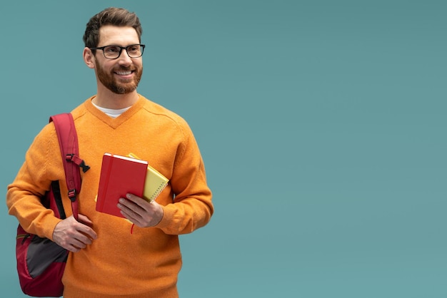 Tevreden jonge student man in casual outfit wegkijken terwijl hij poseert met rugzak en leerboek geïsoleerd op blauwe achtergrond