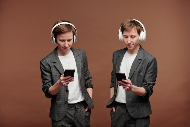 Tevreden jonge tweeling in modieuze pakken die tegen een bruine achtergrond staat en geniet van muziek in een draadloze koptelefoon