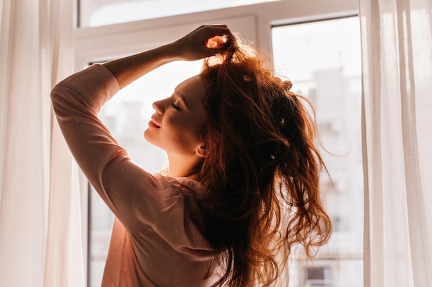 Foto tevreden jonge vrouw die naast het raam poseert binnenopname van een zorgeloos meisje dat speelt met haar lange gemberhaar