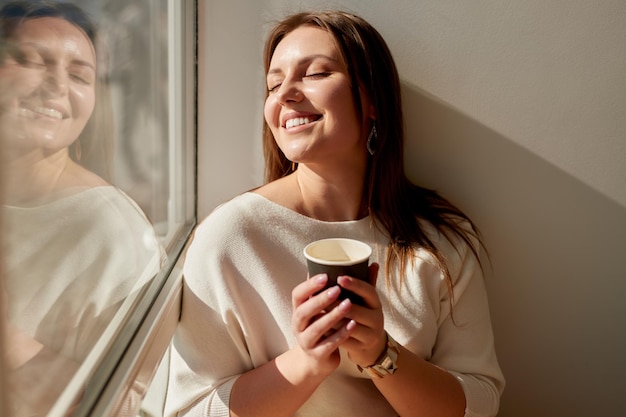 Tevreden jonge vrouw geniet van een zonnige ochtend terwijl ze ontspant bij het raam met een kopje afhaalkoffie thuis