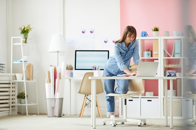 Tevreden jonge vrouw in spijkerkleding die aan tafel op kantoor staat en bestanden controleert op laptop