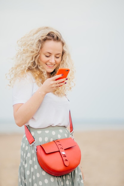 Tevreden jonge vrouw met borstelig krullend haar houdt slimme telefoon maakt gebruik van gratis internetverbinding voor sms-berichten staat tegen wazig zeegezicht achtergrond Mensen en recreatie concept