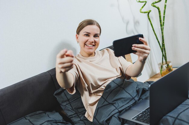 Tevreden jonge vrouw met telefoon die doorwerkt op laptop Vrouw zittend met computer in bed thuis leren op afstand en extern freelance werkconcept