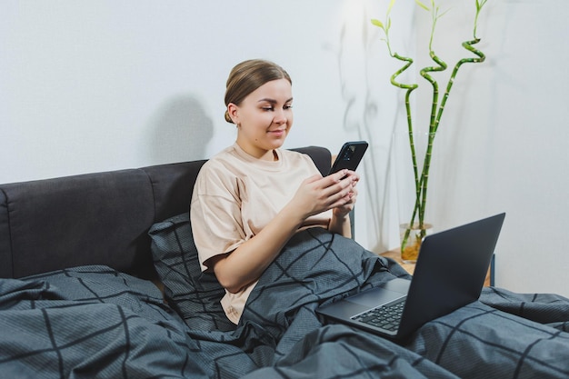 Tevreden jonge vrouw met telefoon die doorwerkt op laptop Vrouw zittend met computer in bed thuis leren op afstand en extern freelance werkconcept