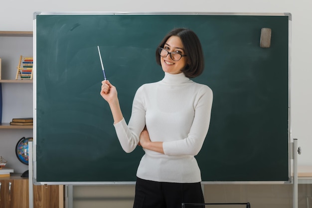 Tevreden jonge vrouwelijke leraar met een bril die voor het bord staat en wijst naar het bord met de aanwijzer in de klas