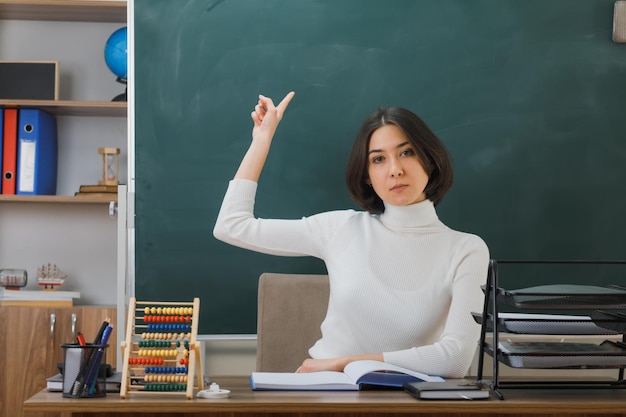 tevreden jonge vrouwelijke leraar wijst naar schoolbord zittend aan een bureau met schoolhulpmiddelen in de klas