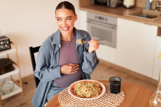 Tevreden jonge zwangere vrouw die huisgemaakte pasta proeft zittend aan tafel in keuken interieur kopie