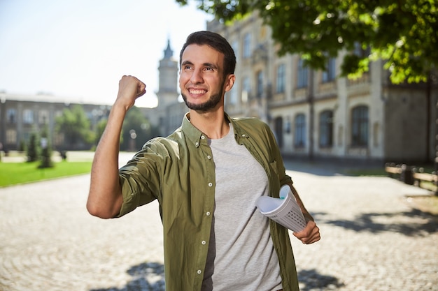 Tevreden jongeman met een werkboek in zijn hand die voor het universiteitsgebouw staat