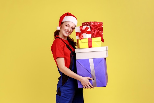 Tevreden koeriersvrouw met ingepakte dozen voor het bezorgen, verzenden van geschenken voor wintervakanties, blauwe overalls en kerstmanhoed dragen. Indoor studio opname geïsoleerd op gele achtergrond.