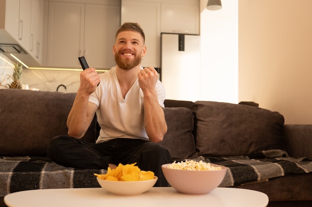 Tevreden man zit op de bank en kijkt tv of film. Jonge Europese bebaarde man met afstandsbediening. Kommen met chips en popcorn op tafel. Concept van thuis rusten. Interieur van studio appartement