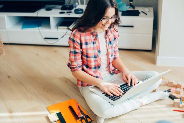 Tevreden meisje in vrijetijdskleding en bril zittend op de vloer met briefpapier aan haar zijde en glimlachend terwijl ze naar het laptopscherm kijkt