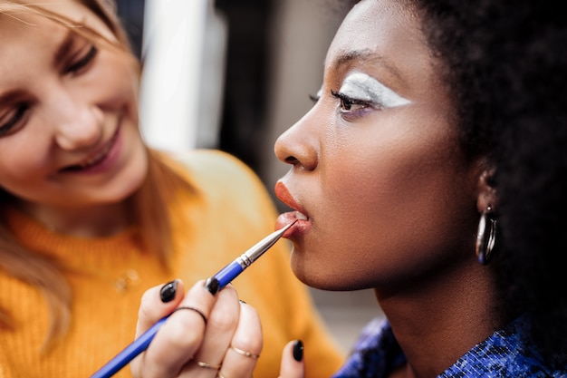 Tevreden met werk. Blonde jonge stylist met zwarte nagellak die mooi lacht terwijl hij lipgloss op de lippen van het model doet