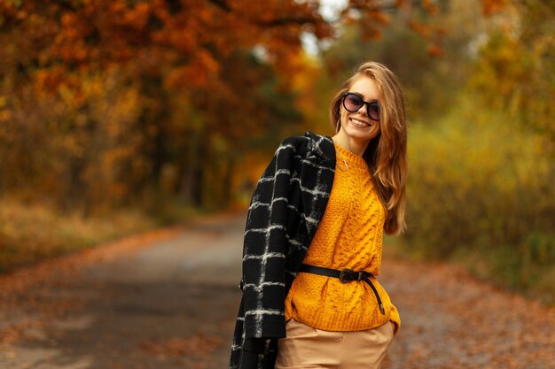 Tevreden mooie jonge slavische vrouw die lacht met haar tanden in een modieuze gele gebreide trui met een jas en zonnebril loopt in een park met oranje herfstbladeren in de natuur