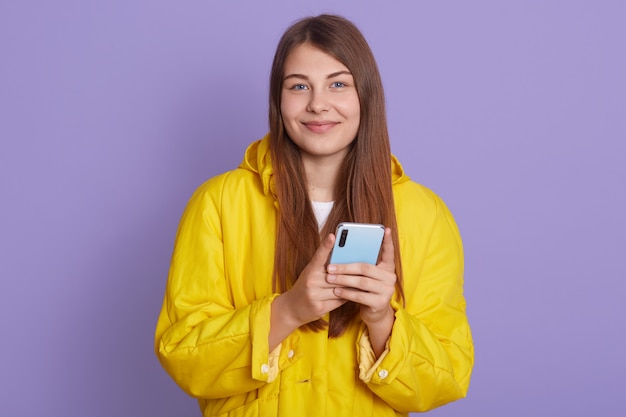 Tevreden mooie vrouw houdt moderne mobiele telefoon, teksten met vriend online in chat, gekleed stijlvol geel shirt staat tegen lila muur.