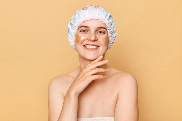 Tevreden opgetogen staande geïsoleerd over beige achtergrond aanraken van haar gezicht aanbrengen van ooglapjes doen huidverzorgingsprocedure na het nemen van een bad in de ochtend
