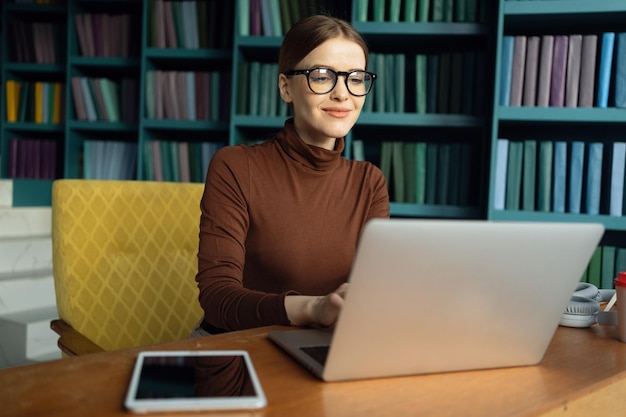 Tevreden professionele vrouw met een bril die aan een laptop werkt in een gezellige bibliotheekomgeving