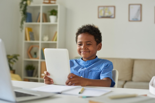 Tevreden schattige Afro-Amerikaanse jongen die aan tafel in de woonkamer zit en tablet gebruikt terwijl hij communiceert