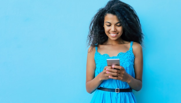 Tevreden tiener Afrikaans etnisch meisje met lang gitzwart haar houdt een smartphone in haar handen, glimlacht terwijl ze op internet surft of feed controleert