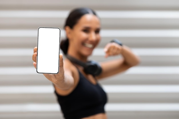 Tevreden vrij millennial arabische vrouwelijke atleet met koptelefoon wijzende vinger naar telefoon met blanco