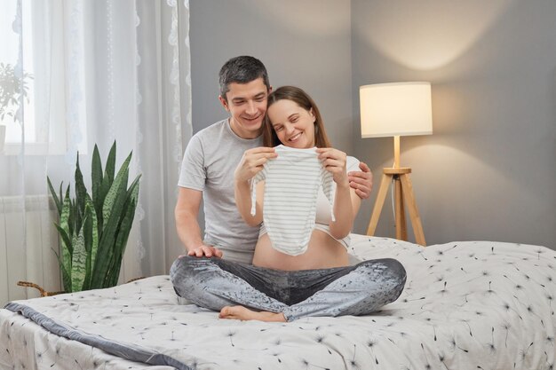 Foto tevreden zwangere vrouw zit op bed met haar man bewondert kleine baby bodysuit wachten op hun dochter of zoon uitdrukking van geluk en positieve emoties