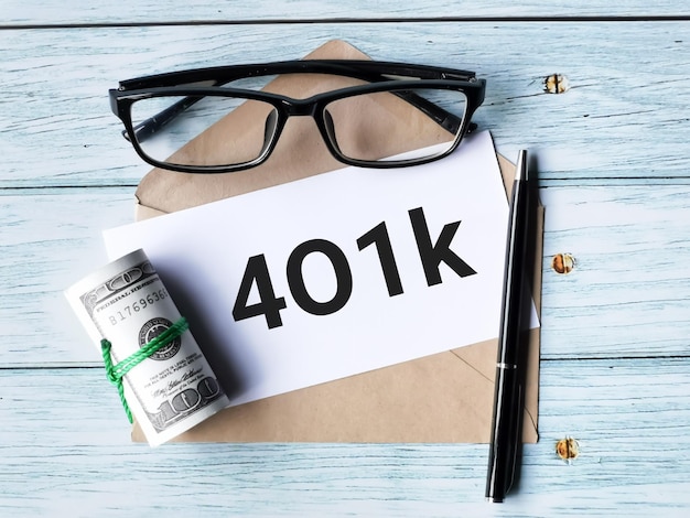 Текст 401k написан на белой бумаге ручкой, коричневым конвертом, фальшивыми деньгами и очками.