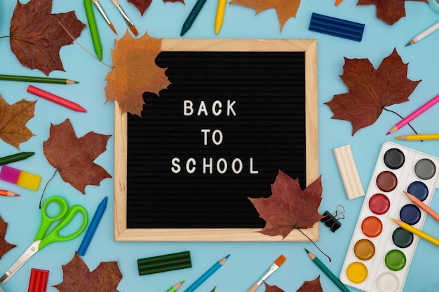 Foto testo ritorno a scuola presto su una lavagna in legno acquisto di cancelleria flatlay sfondo luminoso foglie d'acero tasse scolastiche autunnali